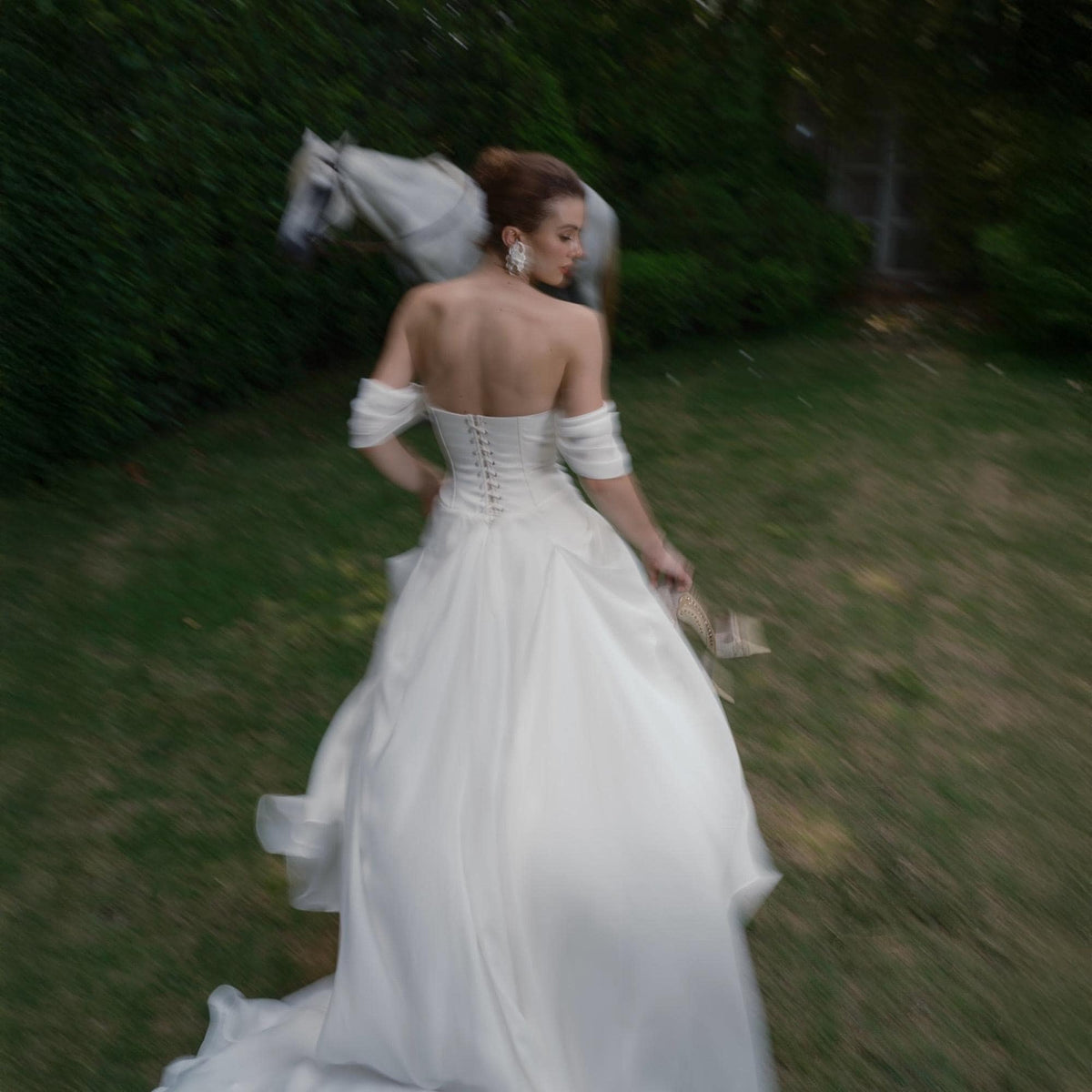 May include: A white strapless wedding dress with a corset back and a flowing skirt. The dress is worn by a person walking on green grass. A white horse is in the background.