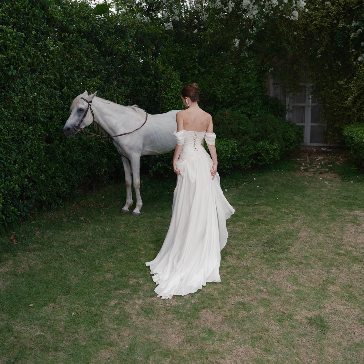 May include: A white horse stands near a person wearing a white off-the-shoulder gown with a flowing skirt. The scene is set in a garden with green grass and foliage, creating a serene and elegant atmosphere. The gown features a corset-style bodice.