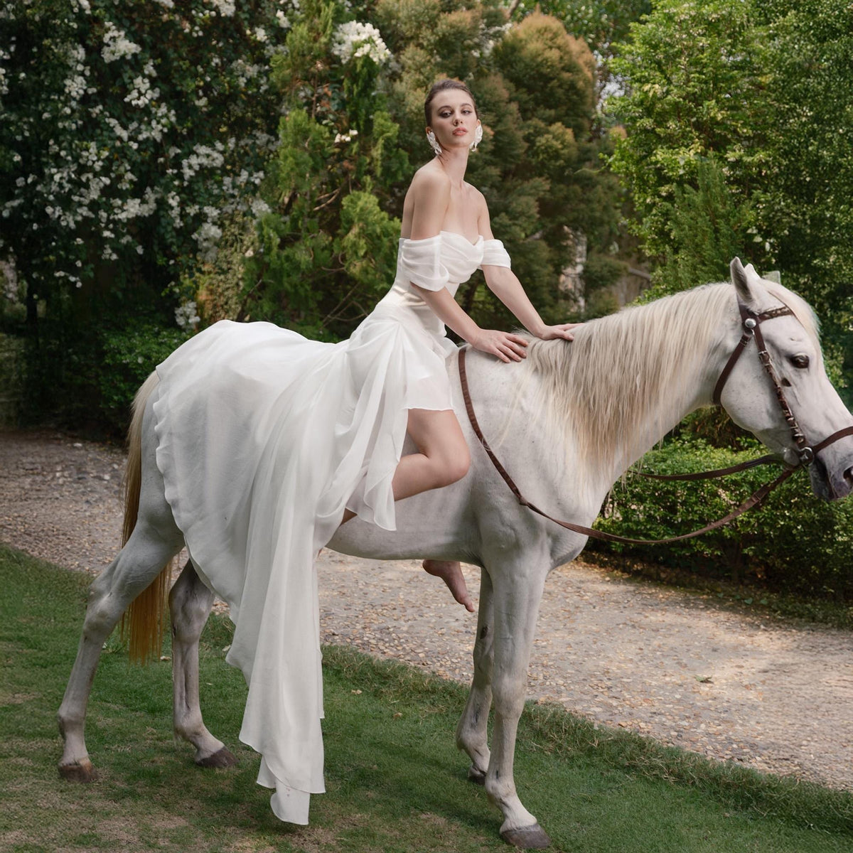 May include: A woman in a white off-the-shoulder dress rides a white horse. The dress has a flowing skirt and short sleeves. The horse has a brown bridle and is on a grassy area with trees in the background.