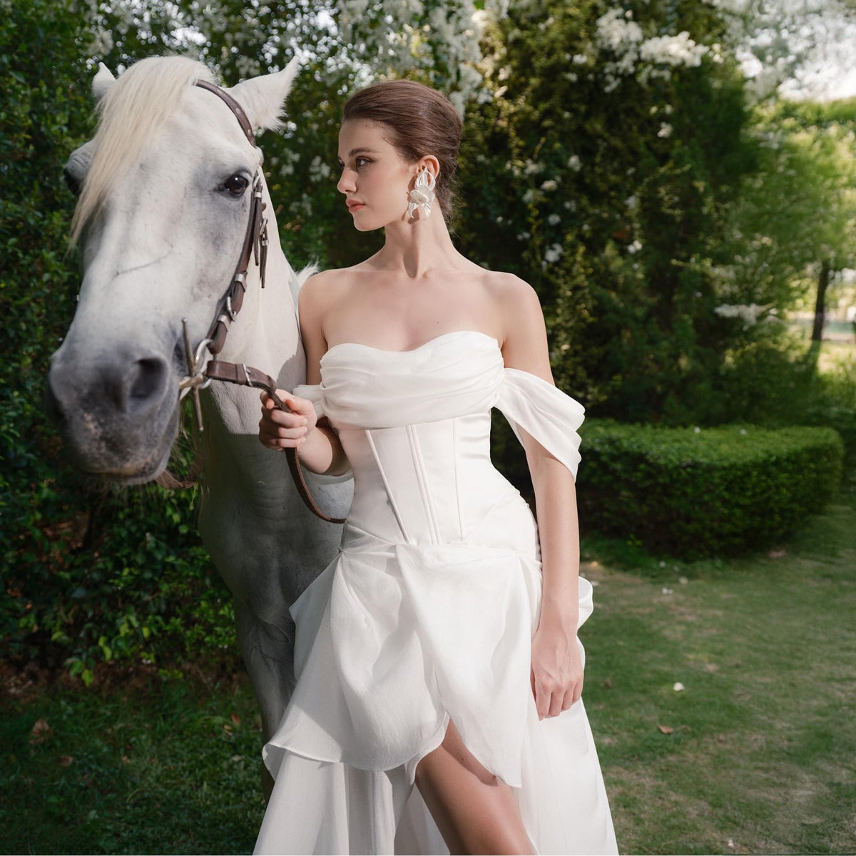 May include: A white strapless wedding dress with a corset bodice and flowing skirt. The dress is paired with large, ornate earrings. A white horse stands beside the dress, with a brown bridle. The scene is set outdoors with greenery in the background.