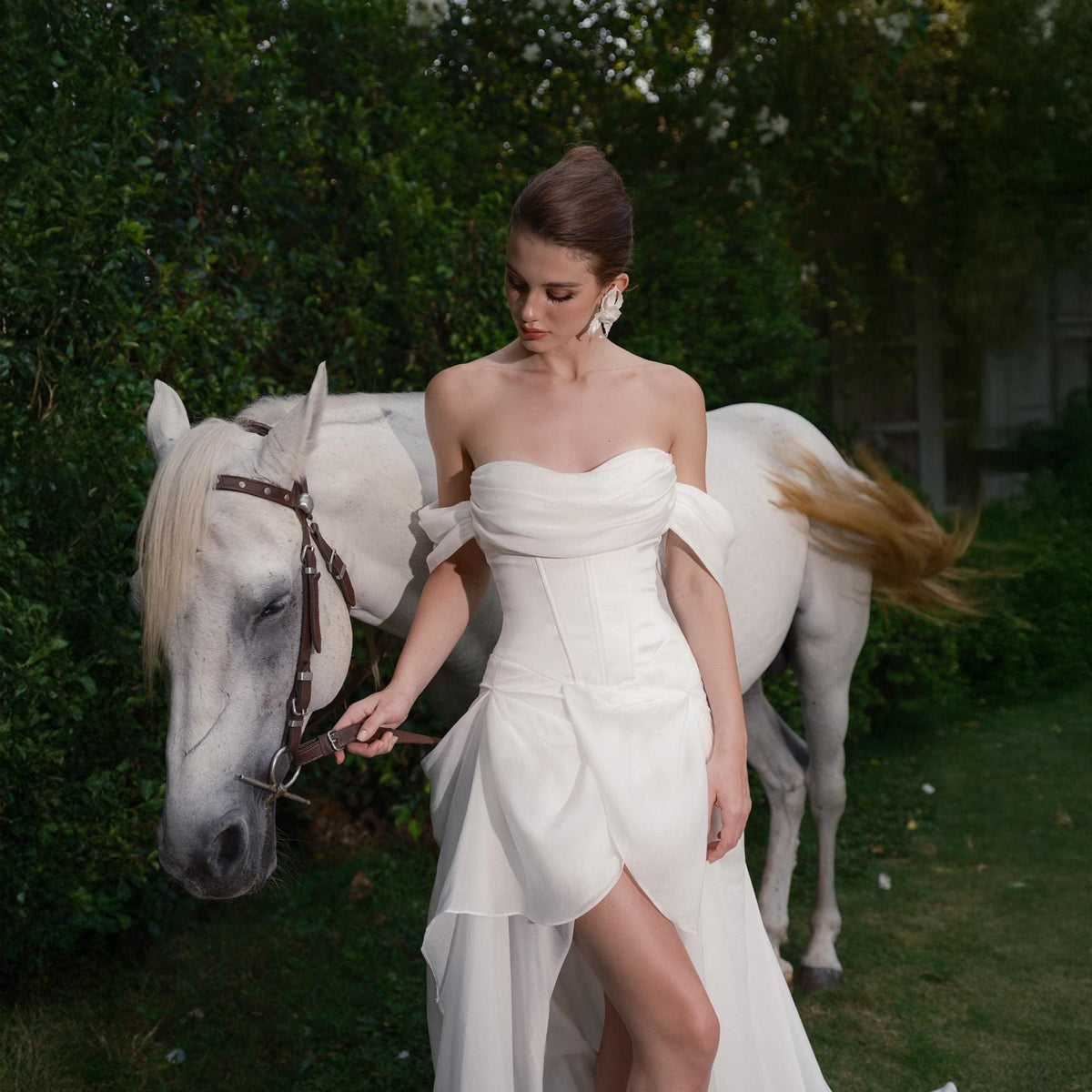 May include: A strapless white wedding dress with a fitted bodice and flowing skirt. The dress features a high slit and is worn outdoors with a white horse in the background. The model is wearing large earrings.