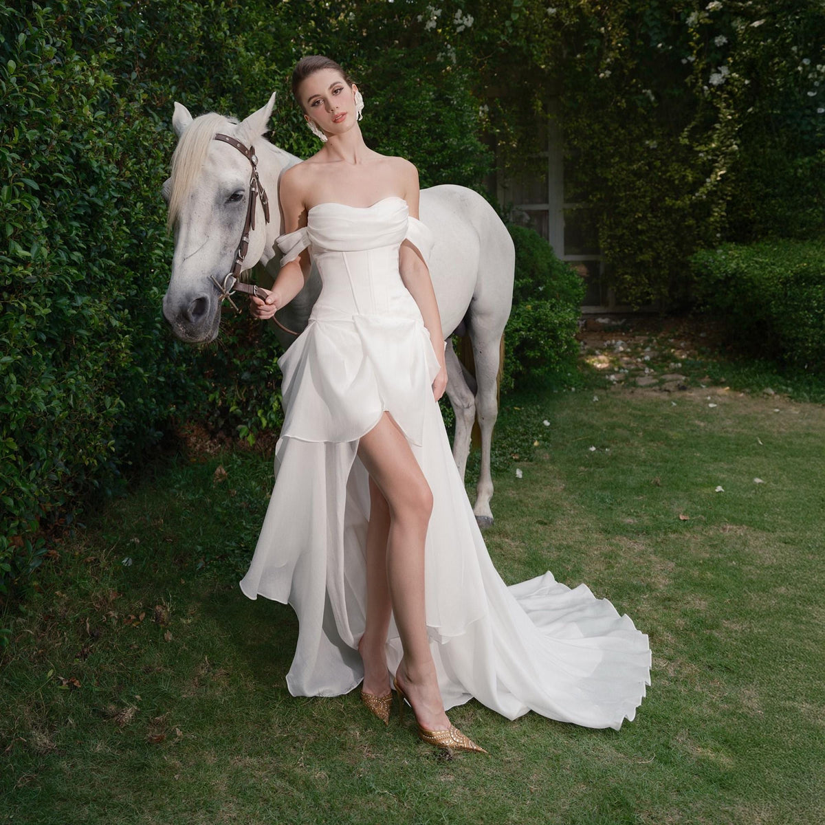 May include: A strapless white wedding dress with a high slit and flowing train. The dress is paired with gold-colored heels. A woman stands next to a white horse in a garden setting.