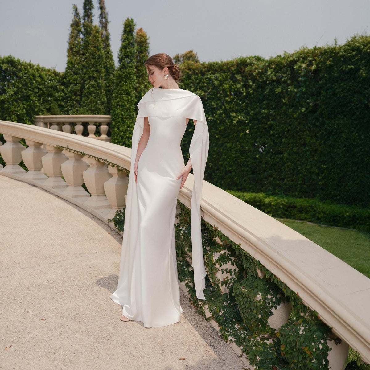 May include: A white, floor-length gown with a draped shawl and long sleeves. The dress is made of a smooth fabric and has a simple, elegant design. The background features a stone balustrade and lush greenery.