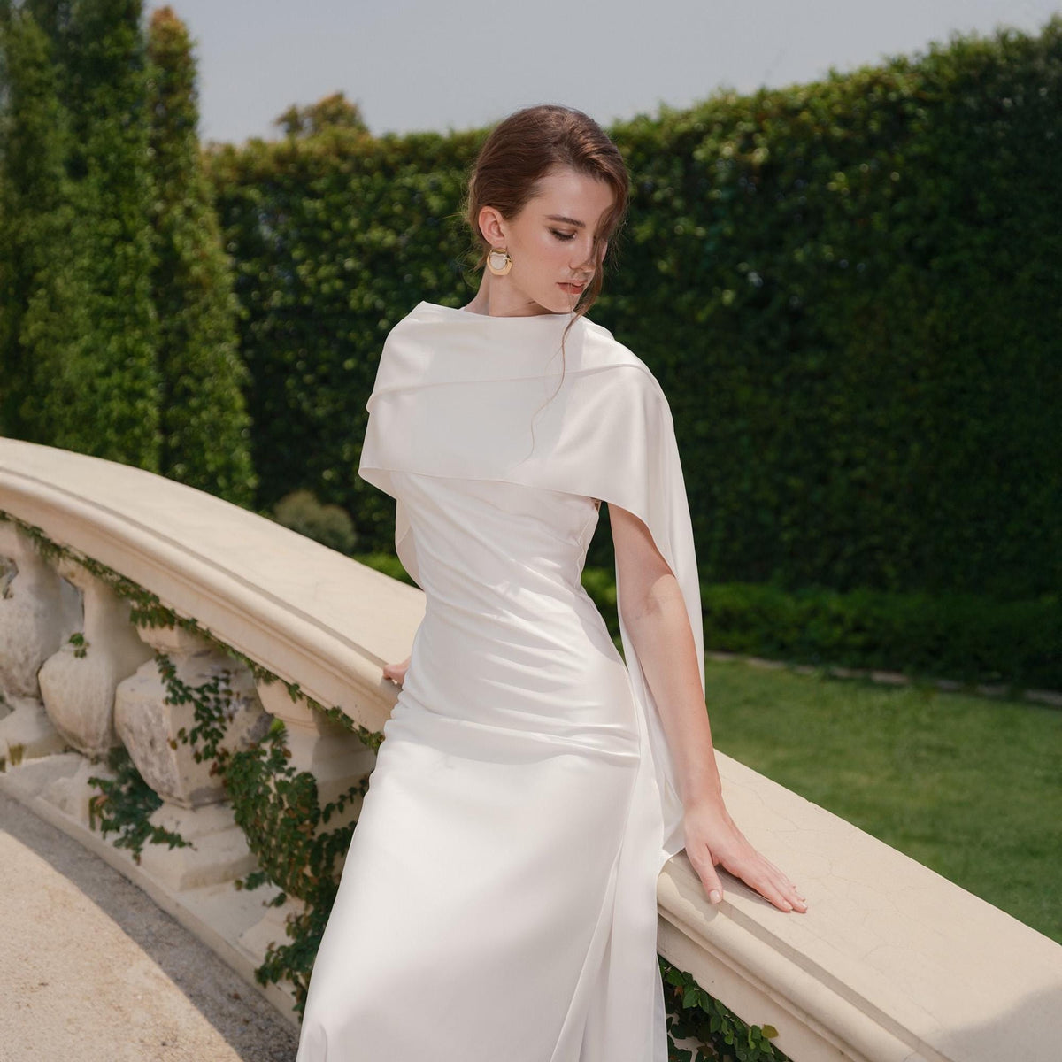May include: A white satin gown with a draped neckline and a flowing skirt. The dress features a cape-like overlay that drapes over the shoulders and arms. The model is standing on a stone bridge with a green hedge in the background.