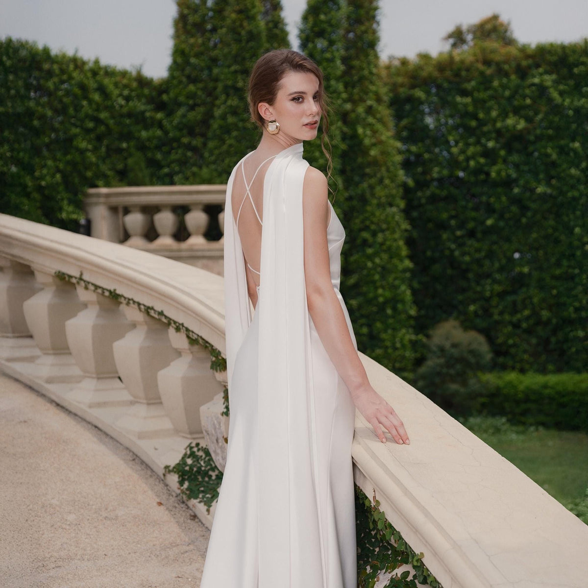 May include: A white, floor-length gown with a high neckline and open back, featuring flowing cape sleeves. The dress is worn outdoors, with a stone balustrade and green foliage in the background.