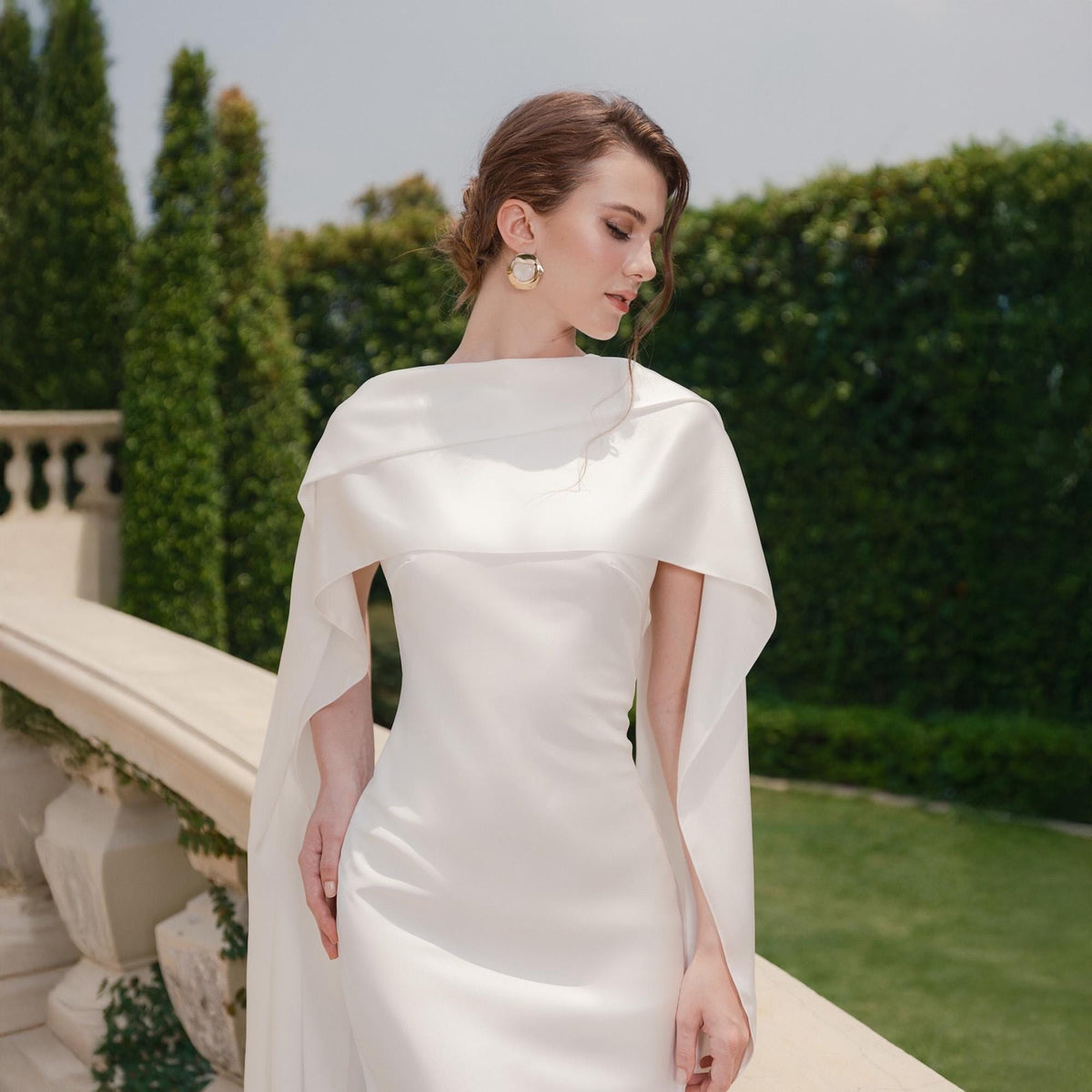 May include: A white wedding dress with a cape-like overlay. The dress has a simple, elegant design with a fitted bodice and a flowing skirt. The cape drapes over the shoulders and arms, creating a graceful silhouette. The background features greenery and a stone railing.