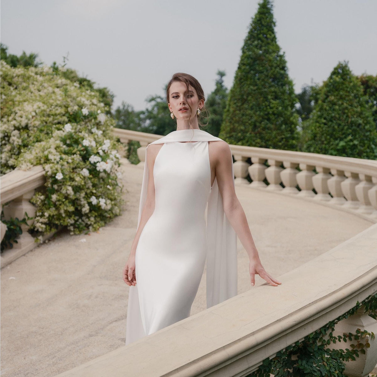 May include: A white, floor-length wedding gown with a high neckline and a flowing cape. The dress is made of a smooth, satin-like fabric. The model is standing on a stone walkway with a stone railing, with greenery in the background.