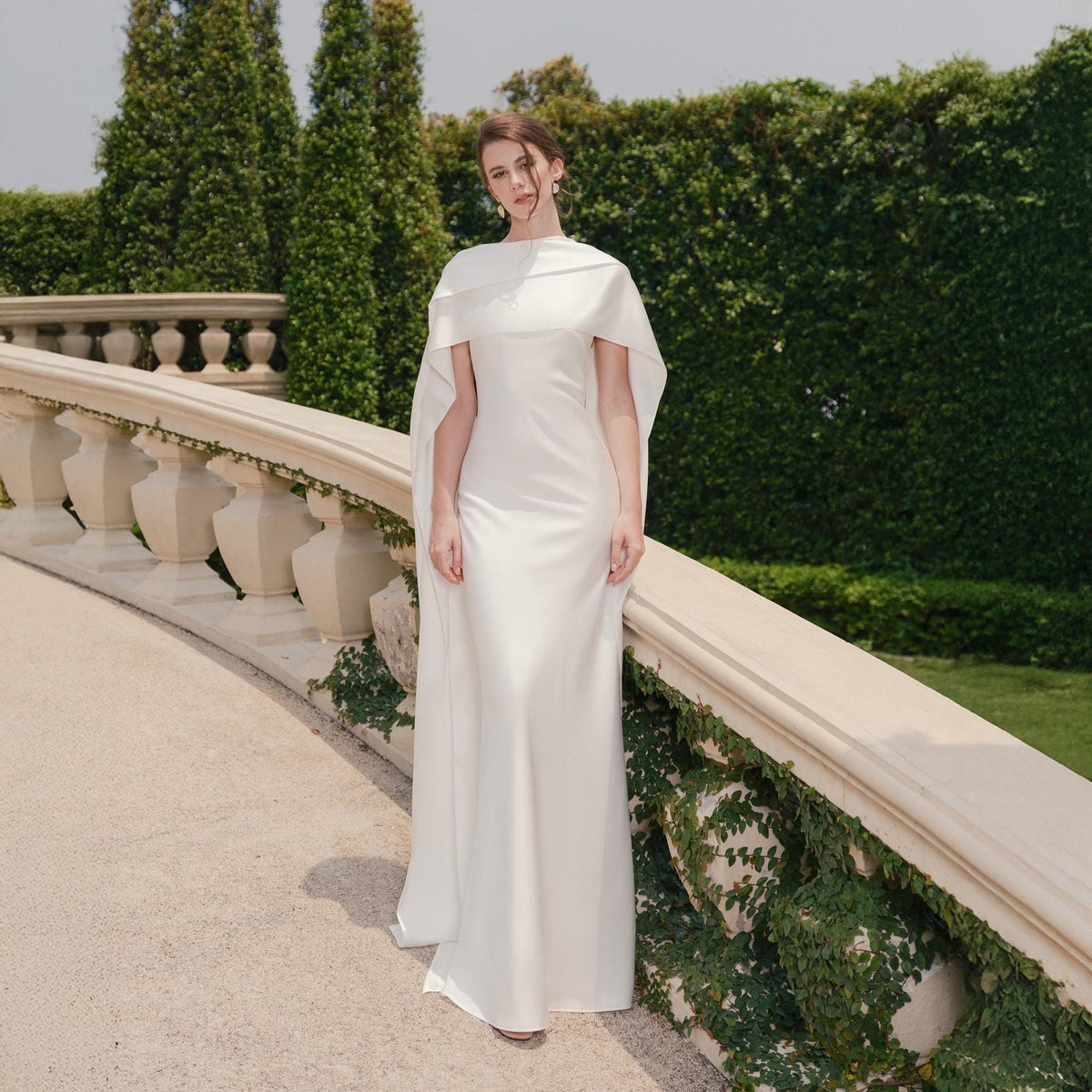 May include: A white, floor-length gown with a cape-like overlay. The dress has a simple, elegant design, with a fitted bodice and a flowing skirt. The cape drapes over the shoulders, adding a touch of sophistication. The background features a stone balustrade and lush greenery.