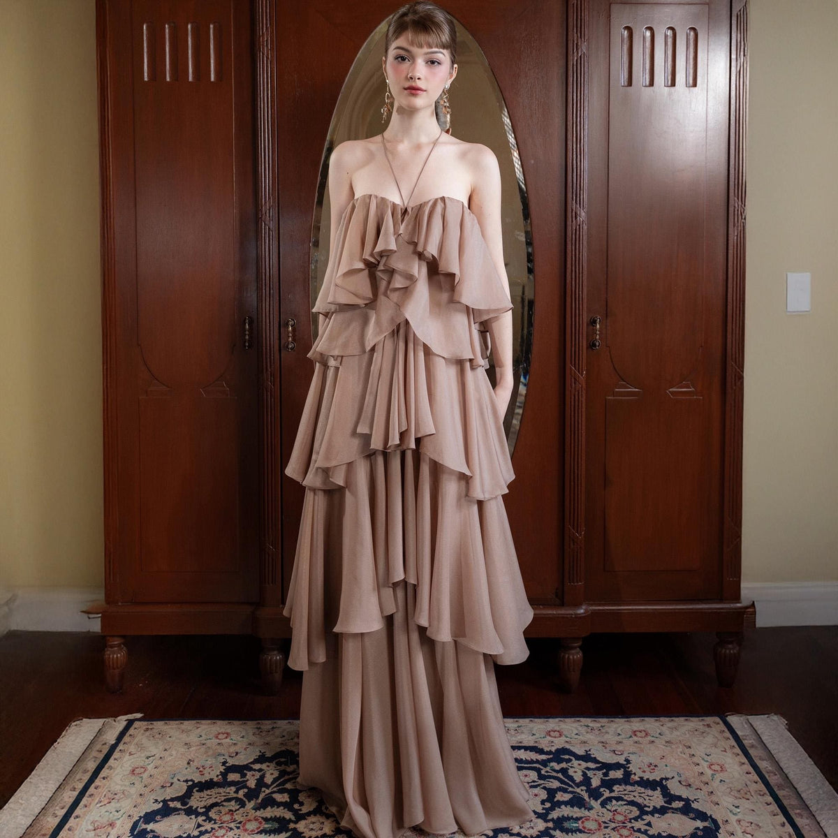 May include: A strapless, tiered, floor-length gown in a soft, neutral beige color. The dress features multiple layers of flowing fabric, creating a ruffled effect. The model is wearing a necklace and earrings. The background includes a wooden wardrobe and a patterned rug.