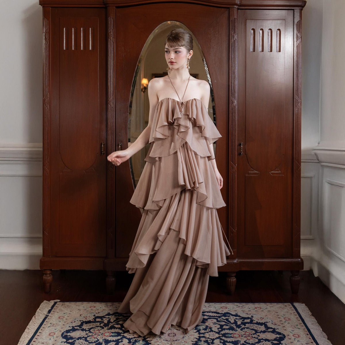 May include: A strapless, floor-length gown in a soft, neutral tone. The dress features multiple layers of flowing fabric, creating a tiered effect. The model is standing in front of a dark wooden wardrobe with a large oval mirror.