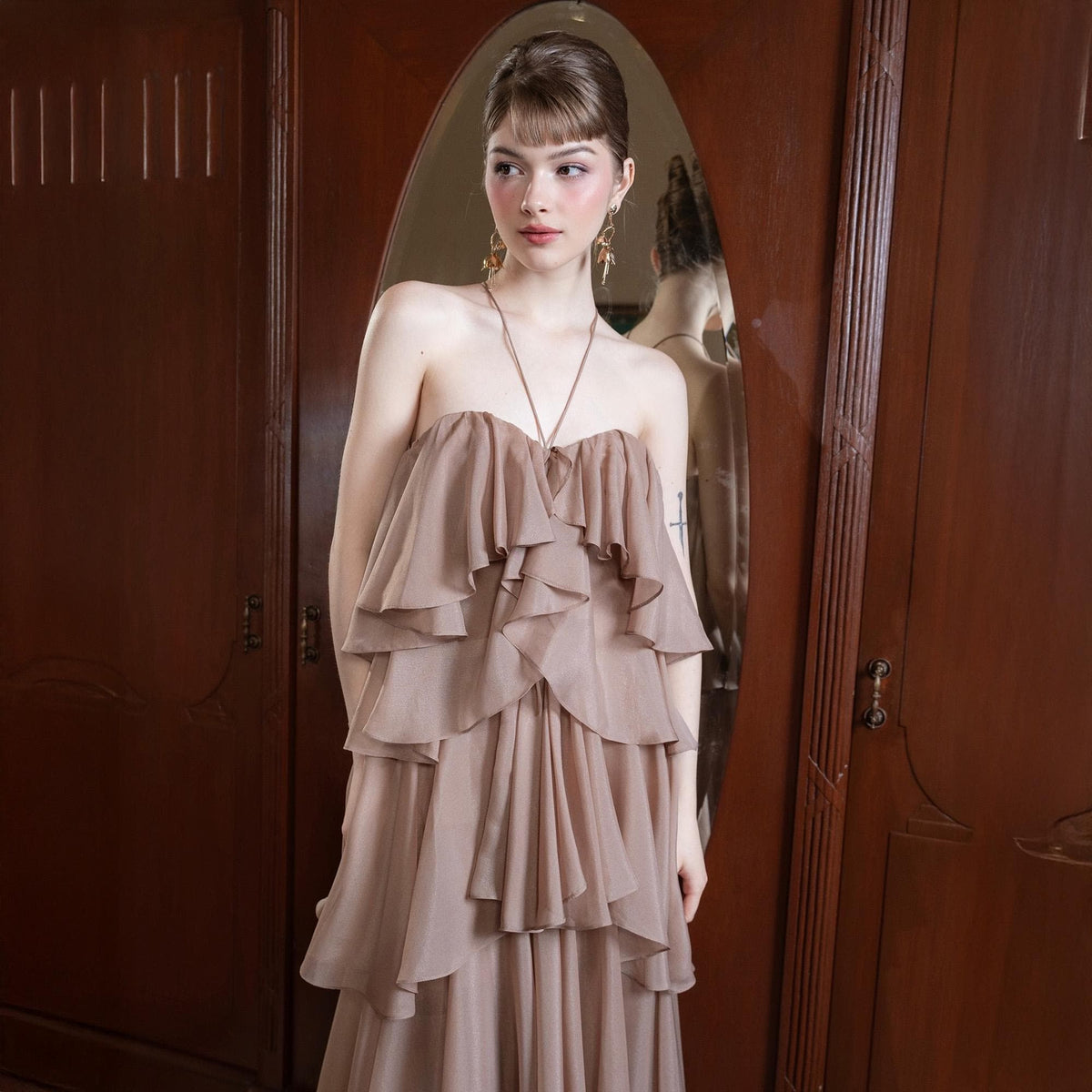 May include: A strapless, layered, taupe-colored dress. The dress features multiple tiers of flowing fabric, creating a ruffled effect. A delicate necklace with a pendant is visible. The dress is suitable for formal events.