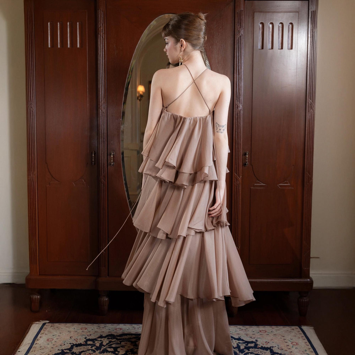 May include: A layered, floor-length dress in a muted mauve color. The dress features a halter-style top with crisscross straps and multiple tiers of flowing fabric. The dress is worn in front of a dark wooden wardrobe with a large oval mirror.