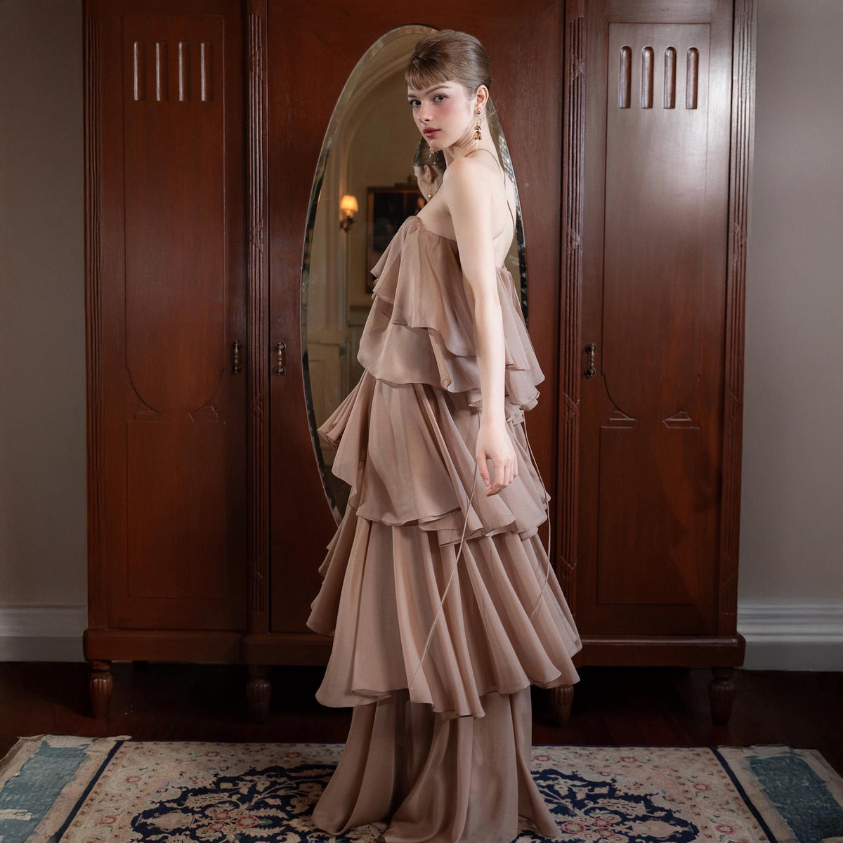 May include: A tiered, floor-length gown in a soft, neutral beige color. The dress features multiple layers of flowing fabric, creating a ruffled effect. The gown has a halter-style neckline and a fitted bodice. The background includes a dark wooden wardrobe and a patterned rug.
