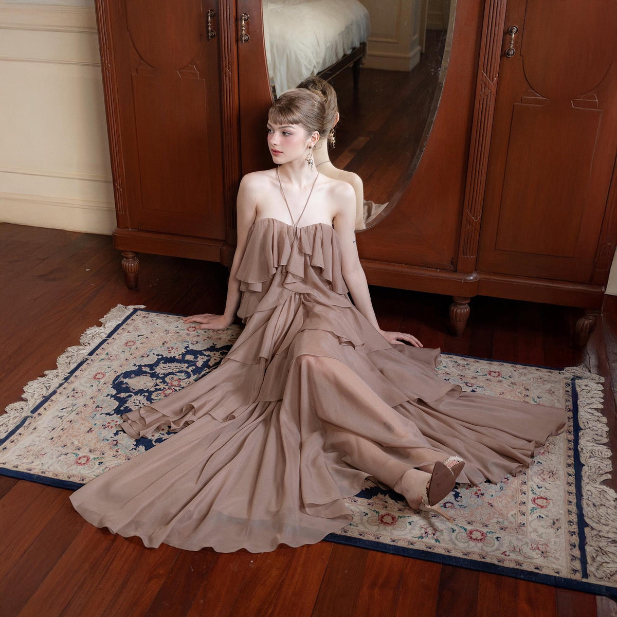 May include: A strapless, floor-length dress in a soft brown hue. The dress features multiple layers of ruffles across the bodice and skirt, creating a flowing, elegant silhouette. The dress is worn on a patterned rug.