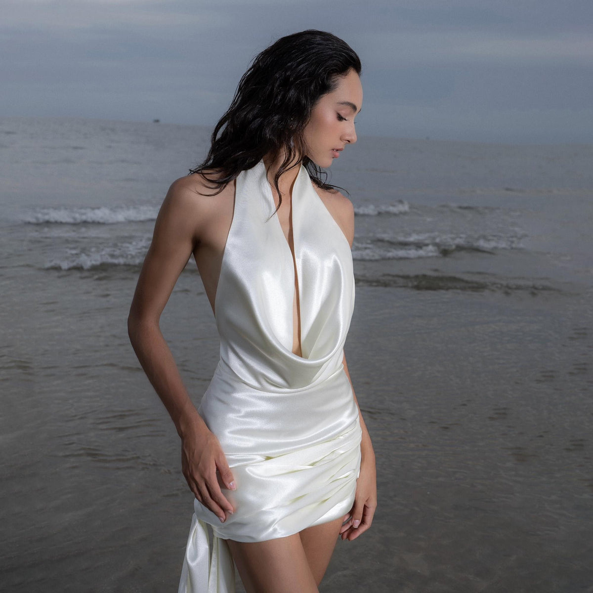 May include: A white satin halter dress with a draped cowl neckline and a short skirt. The dress has a low back and a flowing, gathered detail on the side. The model is standing in the ocean.