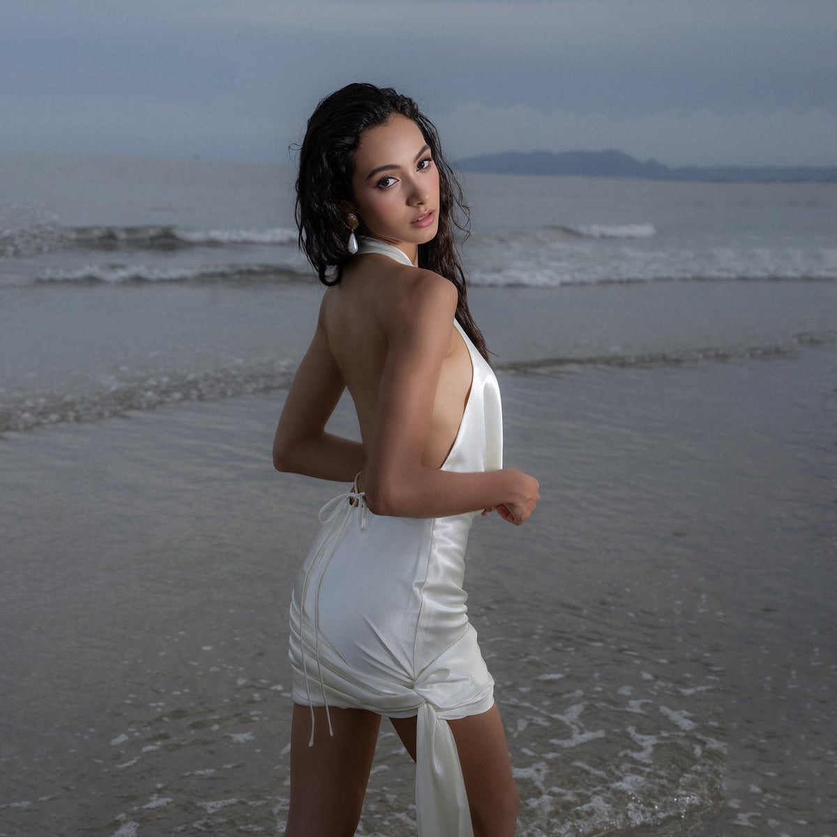 May include: A white satin halter dress with a low back and a draped skirt. The dress is knee-length and has a tie detail at the back. The model is standing in the ocean.