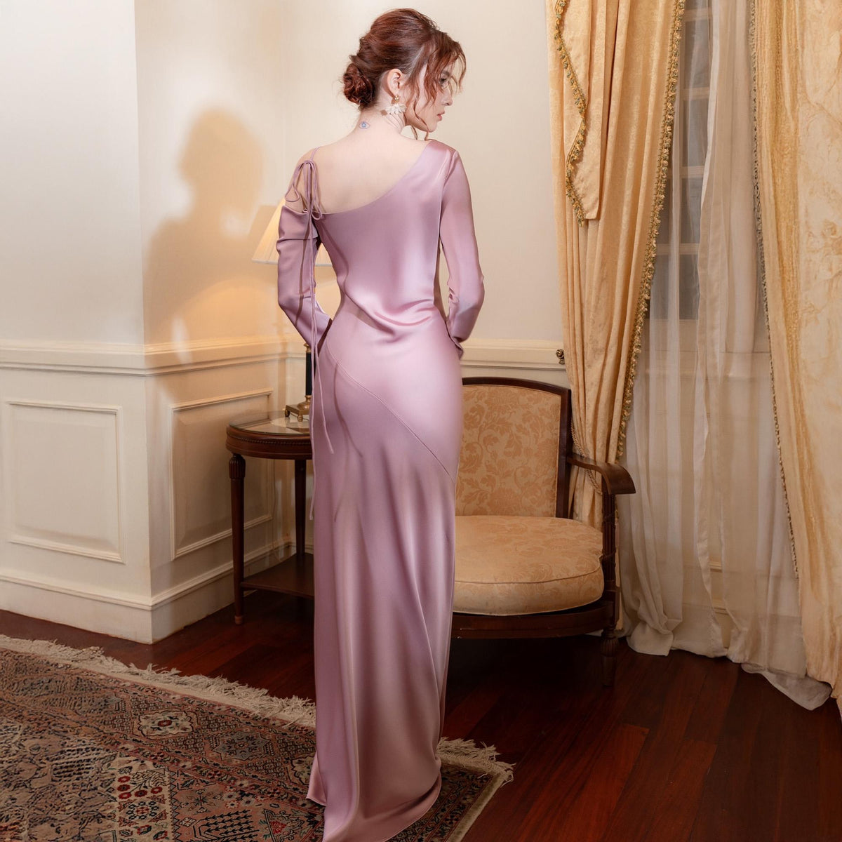 May include: A long, flowing, lilac-colored satin gown with a one-shoulder design and long sleeves. The dress features a diagonal seam across the front and delicate tie details on the shoulder and back. The gown is floor-length.