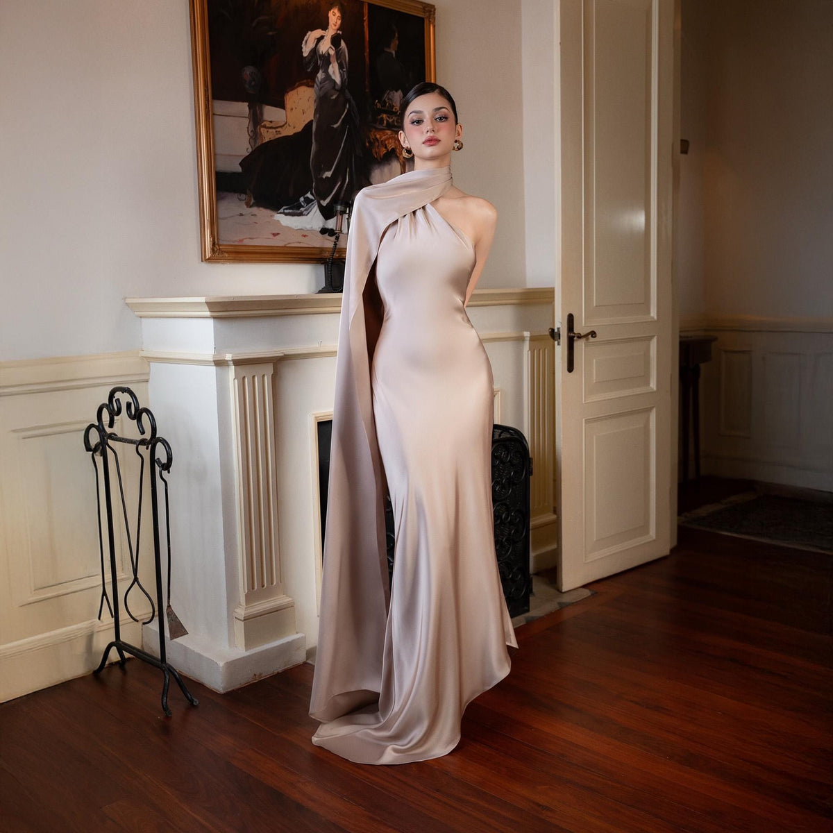 May include: A champagne-colored, one-shoulder evening gown with a flowing cape. The dress has a fitted silhouette that flares out at the bottom. The model is standing in a room with a fireplace and a painting.