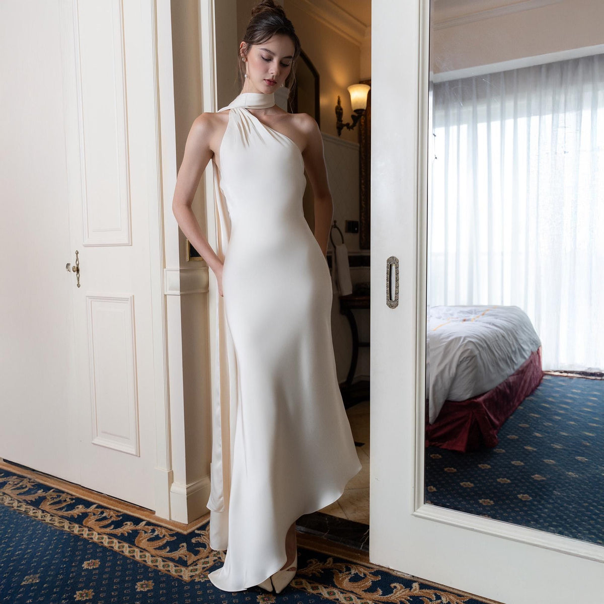 May include: A white, one-shoulder wedding dress with a flowing skirt. The dress features a halter neckline and a long, draped sash. The dress is worn with white heels. The background includes a doorway and a glimpse of a hotel room.