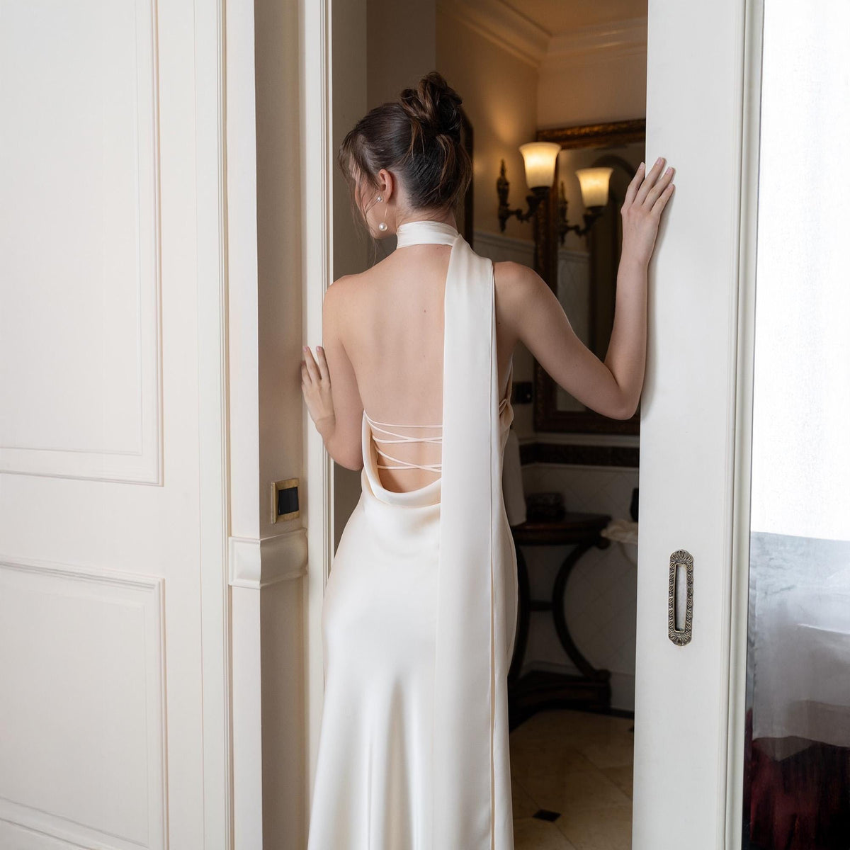 May include: A backless, ivory-colored satin gown with a halter neckline and a flowing, floor-length skirt. The dress features delicate criss-cross straps across the open back. The model is standing in a doorway, with the dress's elegant drape visible.
