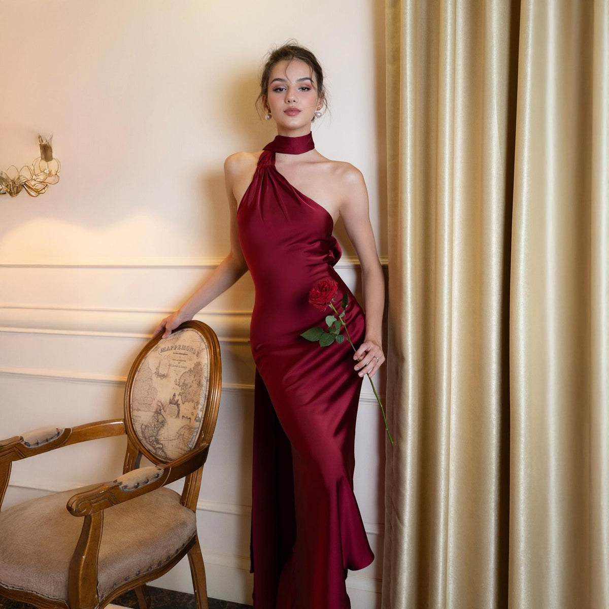 May include: A burgundy satin gown with a halter neckline and a flowing skirt. The dress features a high neck collar and a long, elegant train. A red rose is held in the hand. The background includes a vintage chair and gold curtains.