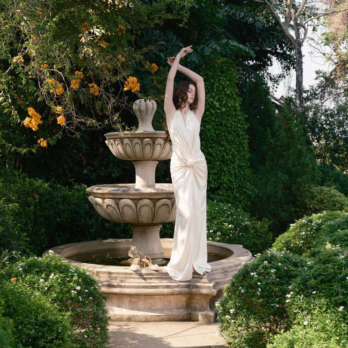 May include: A woman in a white, floor-length dress stands near a tiered fountain in a garden. The dress has a halter neckline and a knot detail at the waist. The fountain is made of stone and surrounded by lush greenery and yellow flowers.