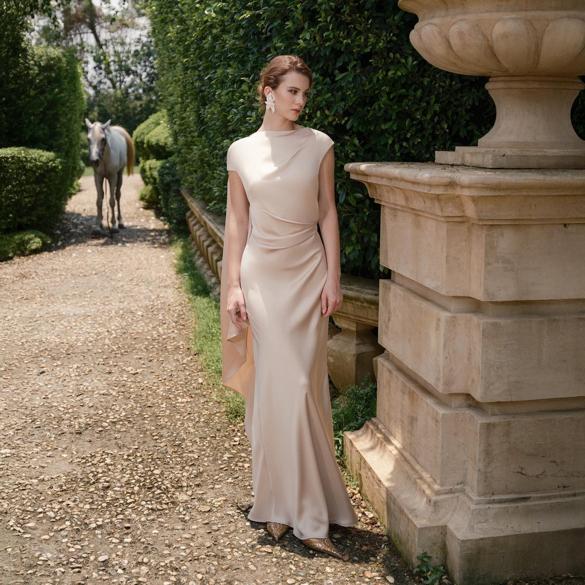 May include: A woman wearing a floor-length, light beige dress with a draped bodice and a flowing skirt. The dress has a subtle sheen and a cape-like detail. The background features a stone pathway, greenery, and a white horse.