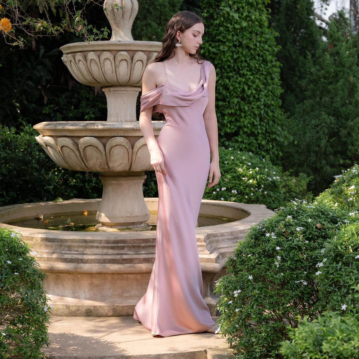 May include: A dusty rose-colored, floor-length gown with a draped neckline and spaghetti straps. The dress has a fitted silhouette and a slight train. The background features a stone fountain and lush greenery, creating an elegant outdoor setting.