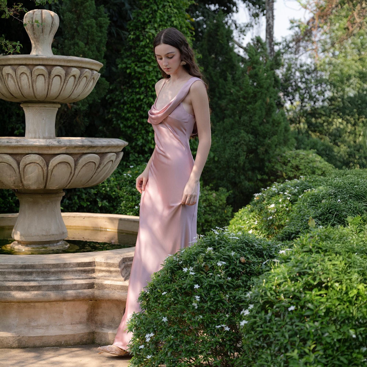May include: A light pink, floor-length slip dress with a cowl neckline. The dress is made of a silky material and has a simple, elegant design. The image is taken outdoors, with a stone fountain and lush greenery in the background.