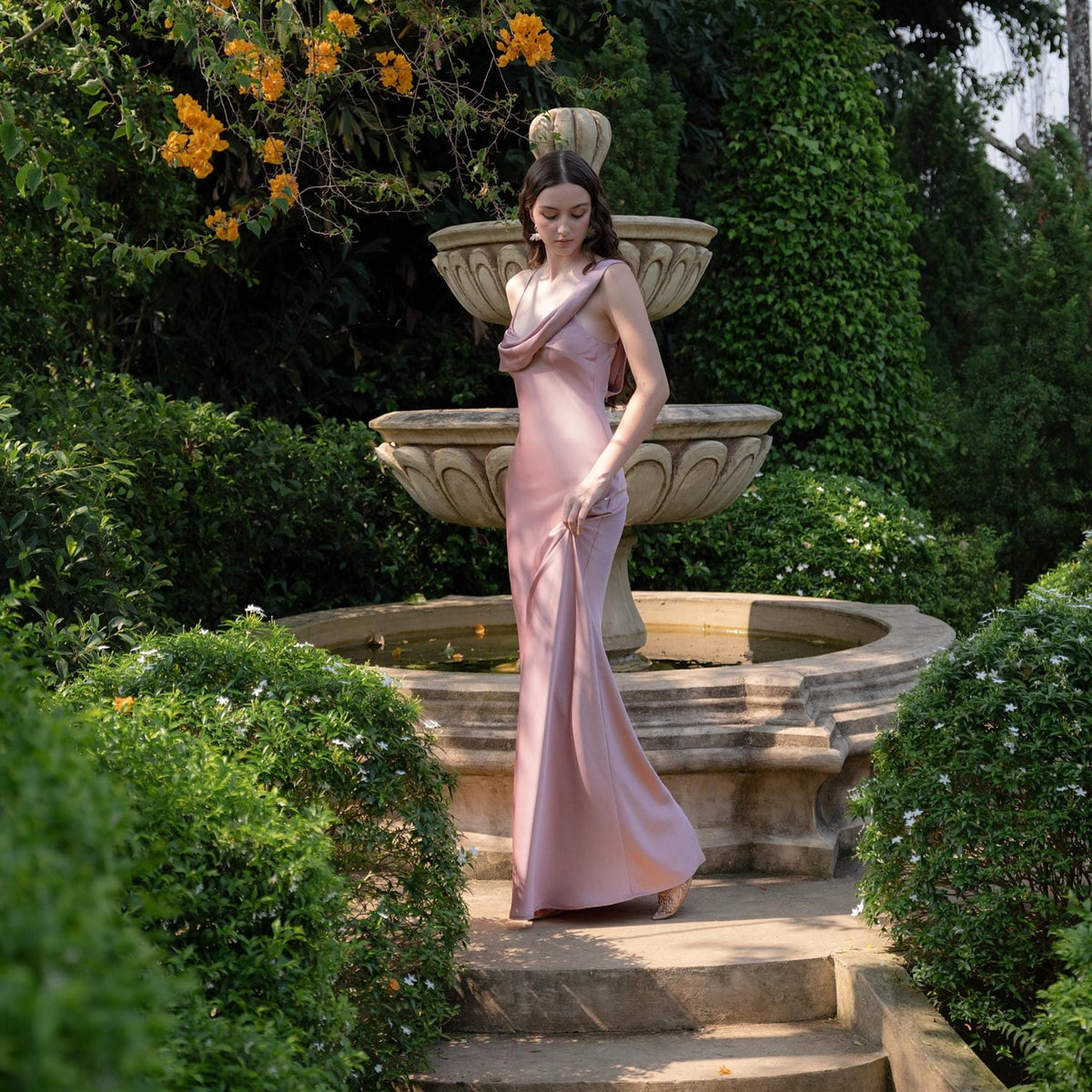 May include: A long, dusty rose-colored satin gown with a cowl neckline and a flowing skirt. The dress is worn outdoors in a garden setting, with a stone fountain and lush greenery in the background.