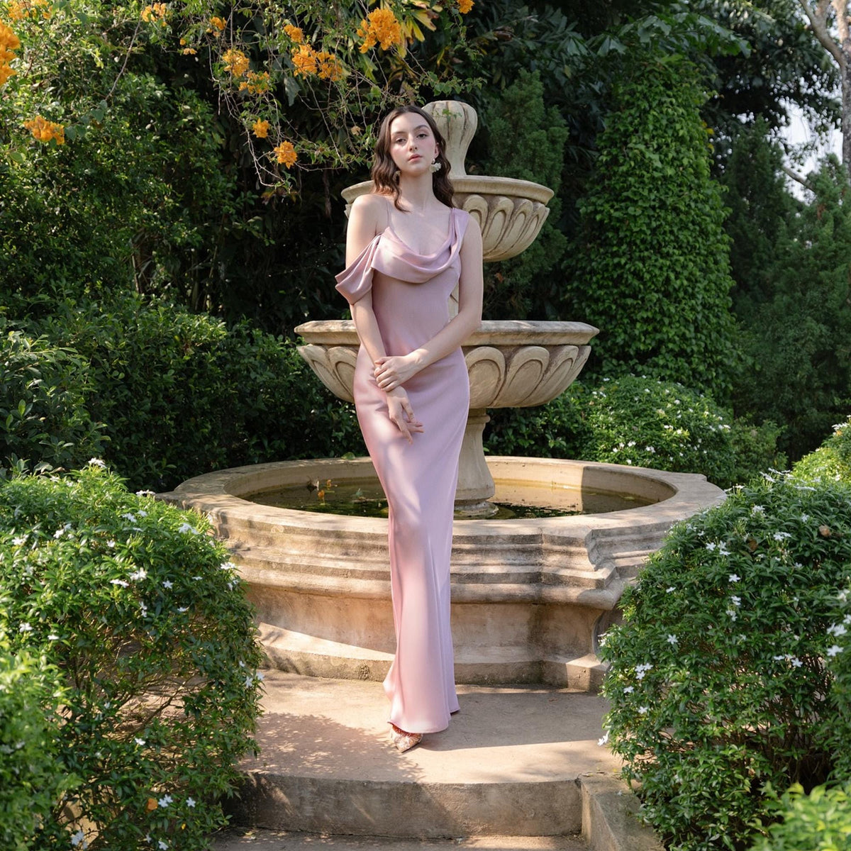 May include: A woman in a light pink, floor-length dress with an asymmetrical neckline stands in a garden. The dress has a draped shoulder detail. A stone fountain and lush greenery are in the background.