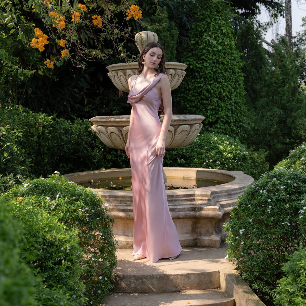 May include: A light pink, floor-length dress with a cowl neckline. The dress is made of a silky material and is worn outdoors near a tiered fountain. The background includes lush greenery and yellow flowers.