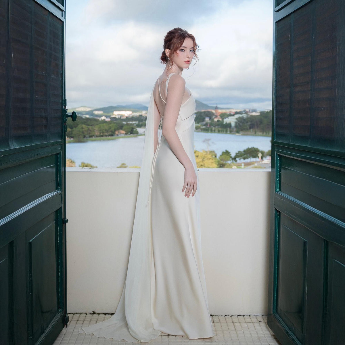 May include: A cream-colored, floor-length gown with spaghetti straps and a flowing chiffon cape. The dress has a simple, elegant design. The model is standing on a balcony with a scenic view of a lake and mountains.
