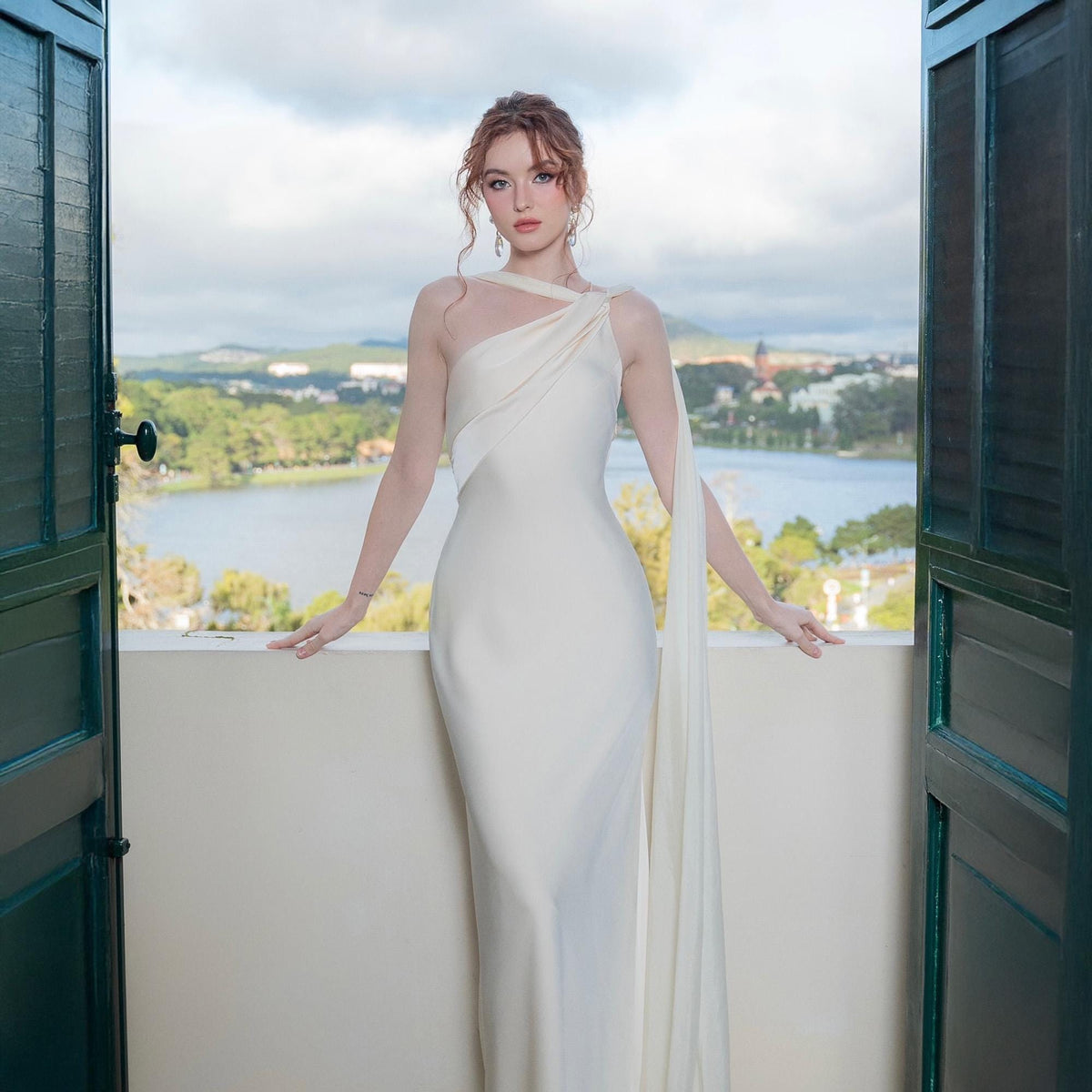 May include: A white, one-shoulder gown with a flowing, draped design. The dress features a fitted silhouette and a long, elegant train. The model is standing on a balcony with a scenic view of a lake and mountains in the background.