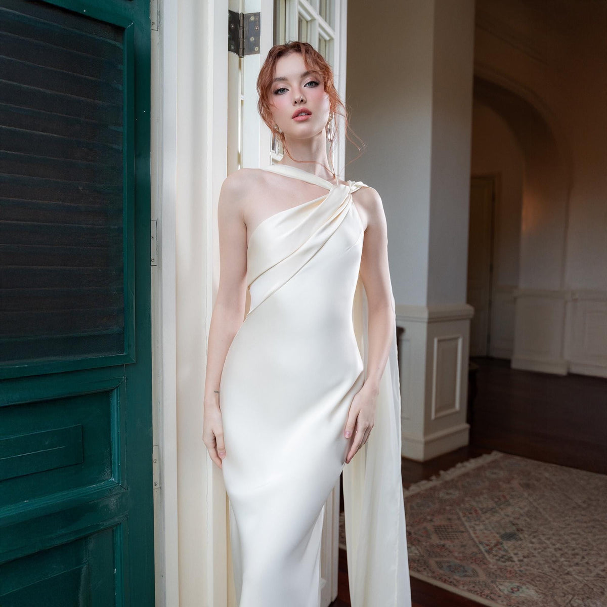 May include: An ivory-colored, one-shoulder gown with a draped neckline and a flowing cape. The dress is floor-length and form-fitting, made of a smooth, satin-like fabric. The model is standing in a doorway with a green door and a patterned rug in the background.
