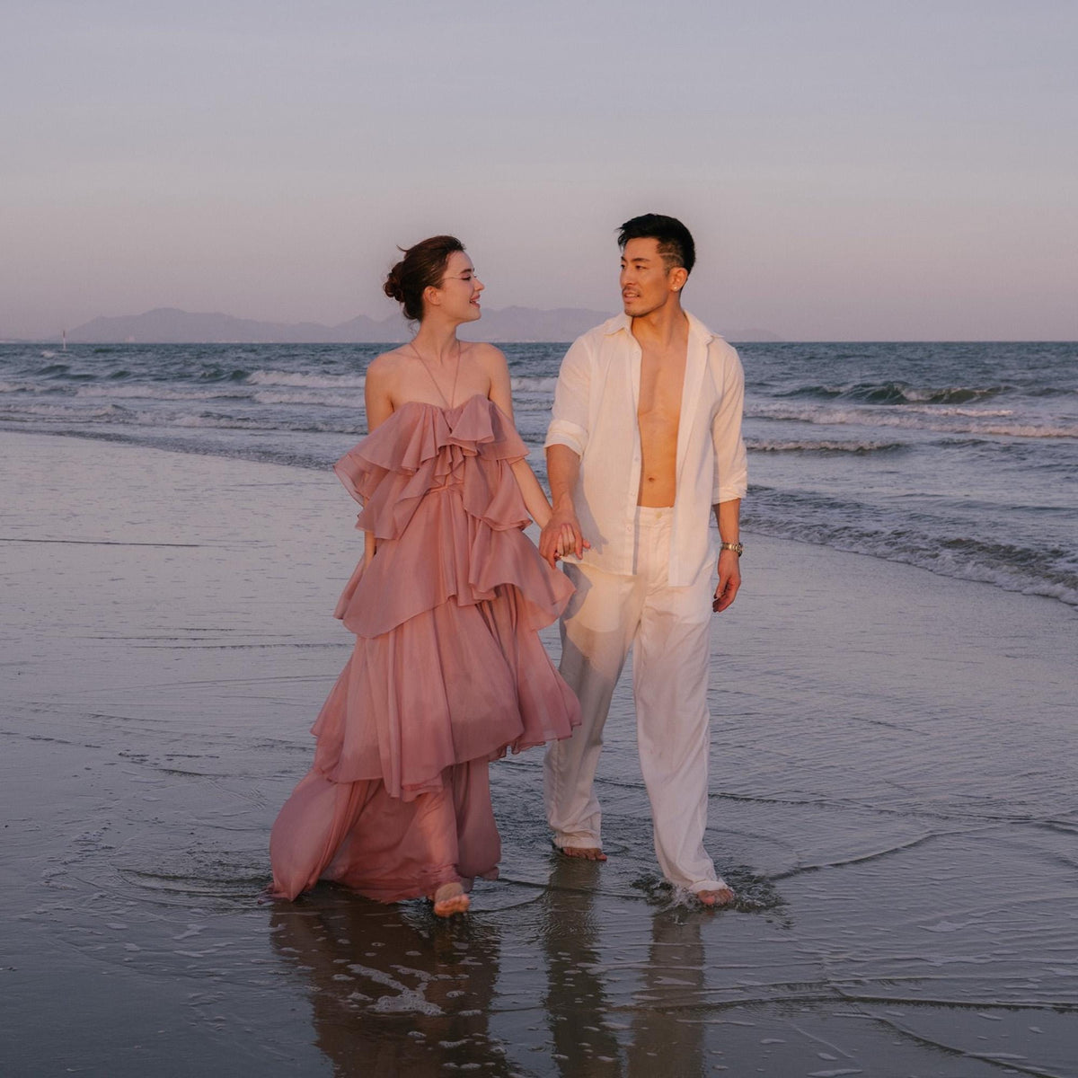 May include: A couple walks hand-in-hand in the ocean. The woman wears a layered, dusty rose-colored gown. The man wears a white button-down shirt and white pants. The sky is a soft pink and blue, and the water is calm.