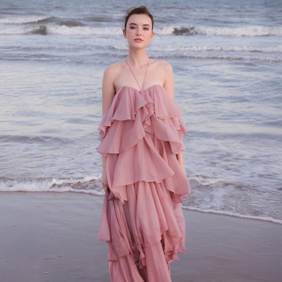 May include: A strapless, layered, dusty rose dress. The dress features multiple tiers of flowing fabric, creating a ruffled effect. A silver necklace hangs from the neck. The background is a beach with the ocean in the distance.