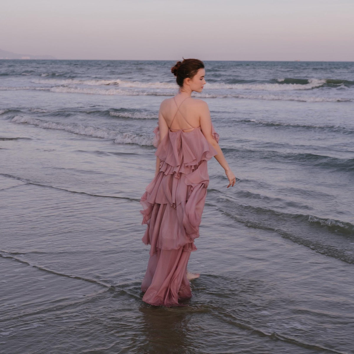 May include: A dusty rose, tiered maxi dress with spaghetti straps and a flowing silhouette. The dress is worn by a person standing in the ocean, with the sea and sky in the background. The dress has a layered, ruffled design.