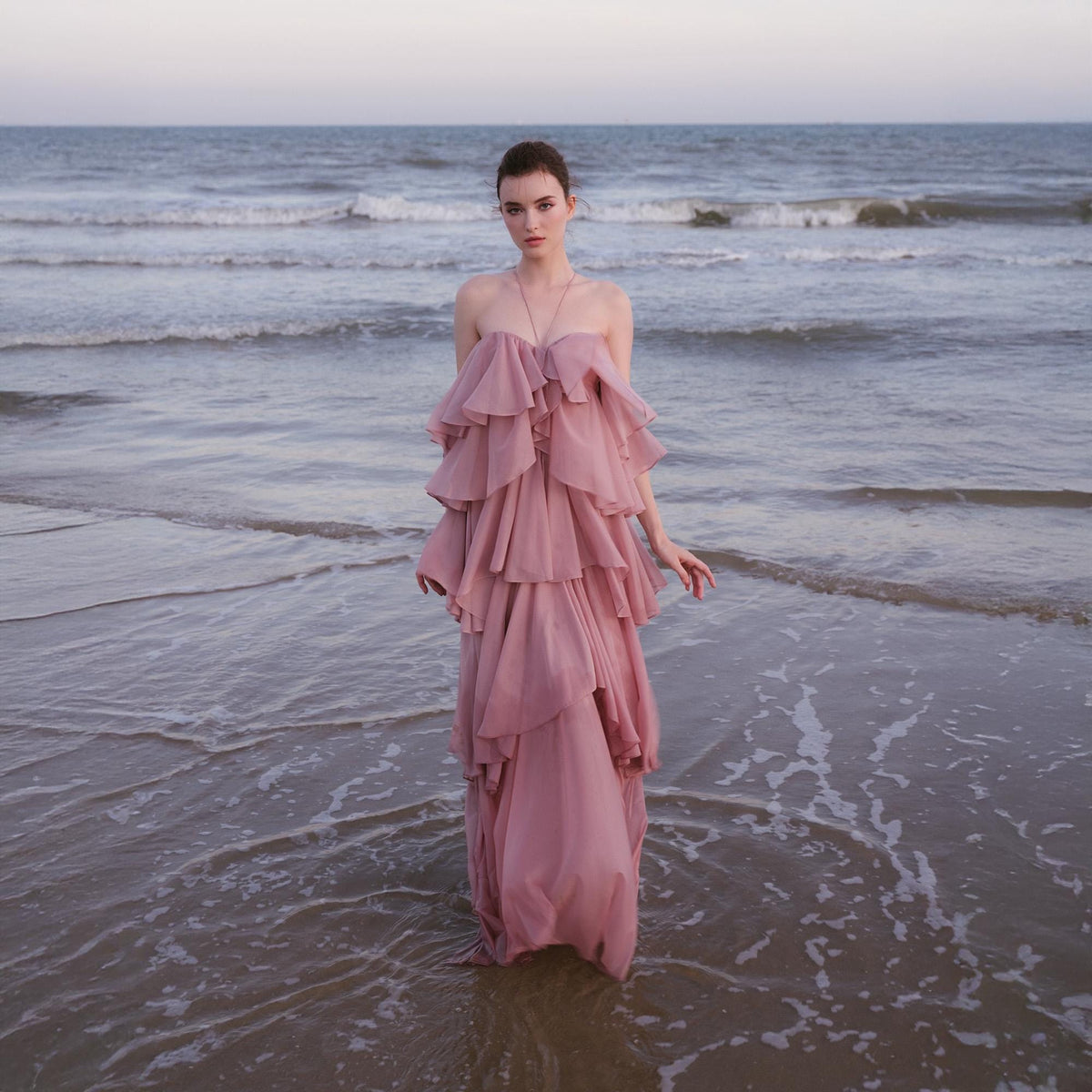 May include: A dusty rose, strapless gown with layered ruffles. The dress is floor-length and worn by a person standing in shallow water at the beach. The ocean and sky are visible in the background.