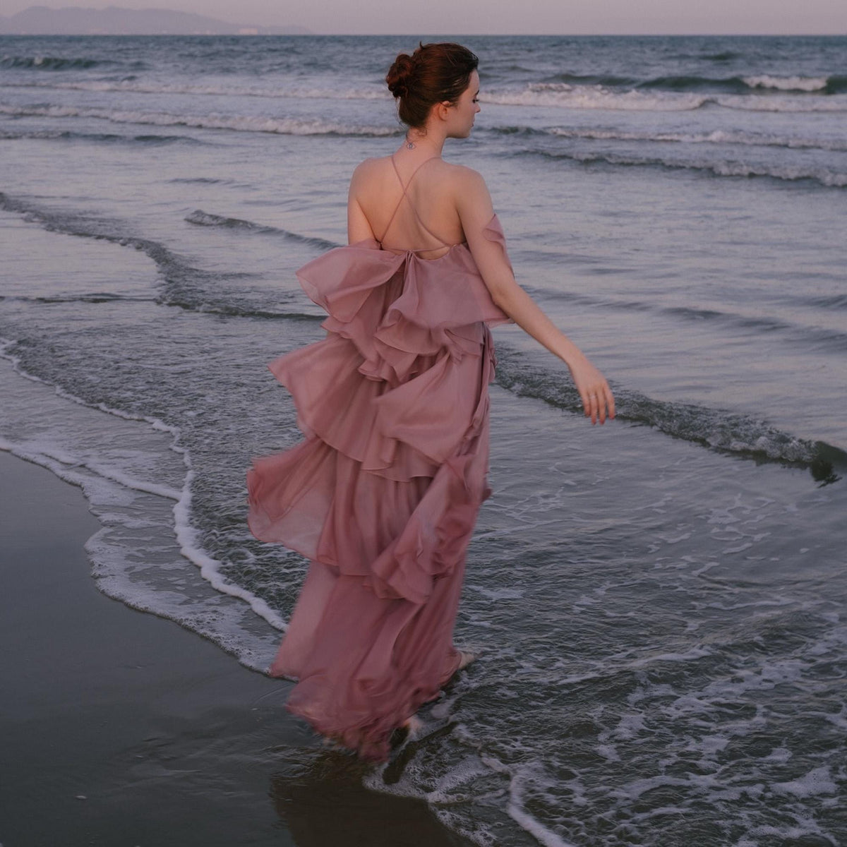May include: A flowing, dusty rose-colored dress with layered ruffles. The dress has thin straps and a low back. The person is standing in the ocean, with the waves gently lapping at the shore. The sky is a soft, muted color.