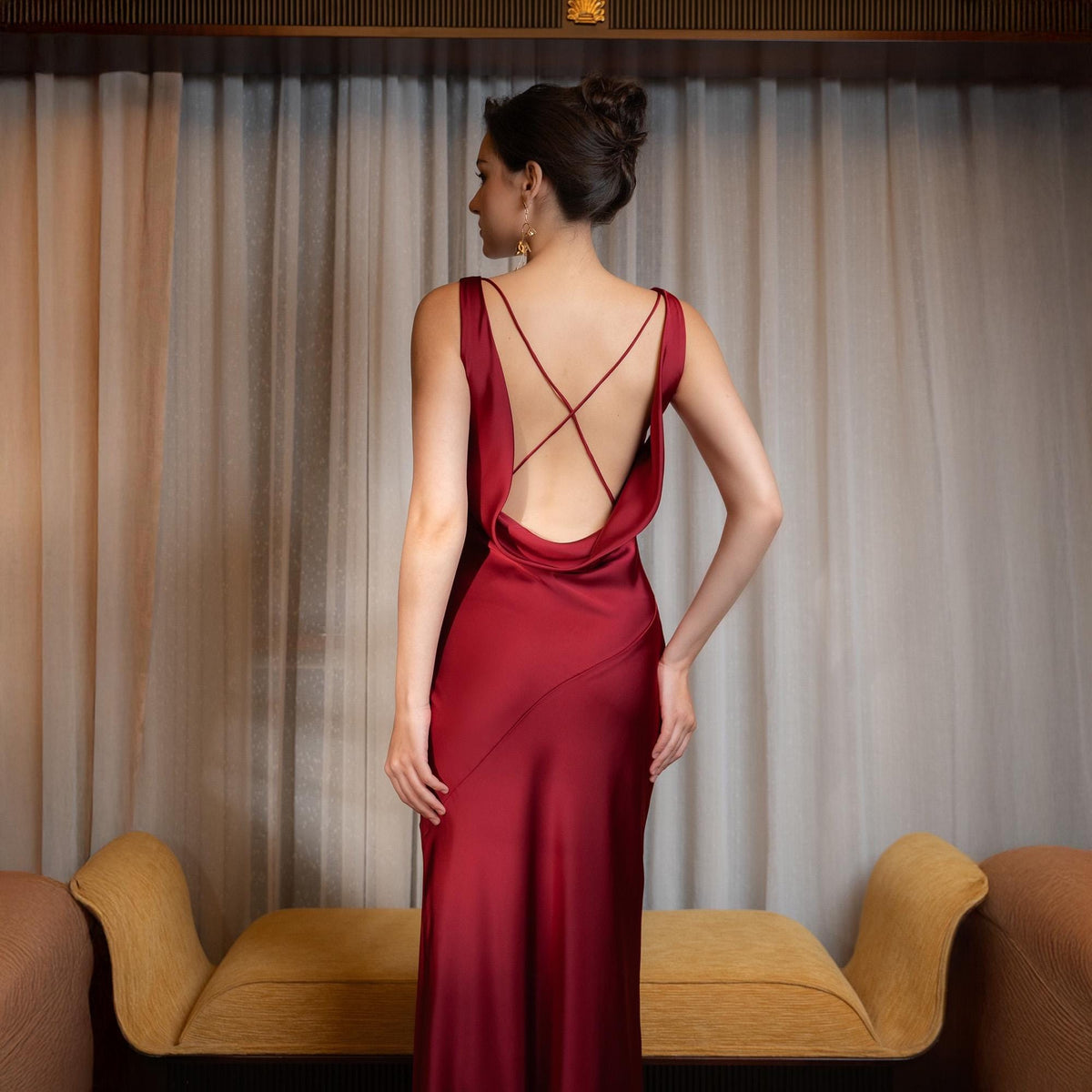 May include: A burgundy-colored, floor-length dress with a low, open back and spaghetti straps that cross. The dress has a draped cowl neckline and a subtle sheen. The model is standing in front of a neutral-toned curtain and a yellow sofa.