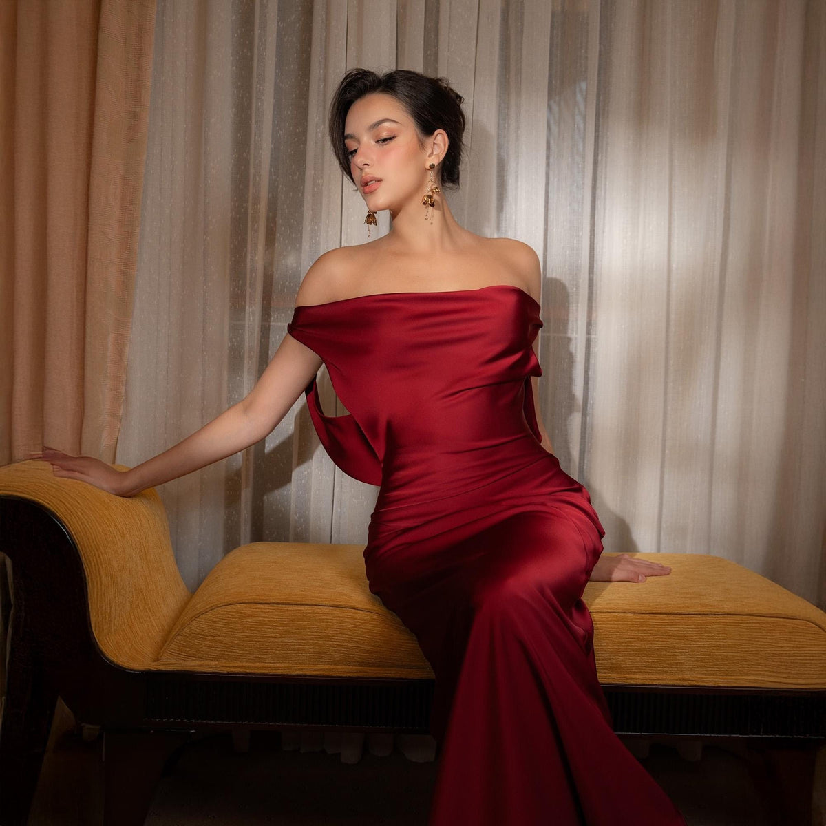 May include: A deep red, off-the-shoulder satin gown. The dress has a flowing silhouette and is paired with gold-toned earrings. The model is seated on a yellow bench with dark wood trim, against a backdrop of neutral-toned curtains.