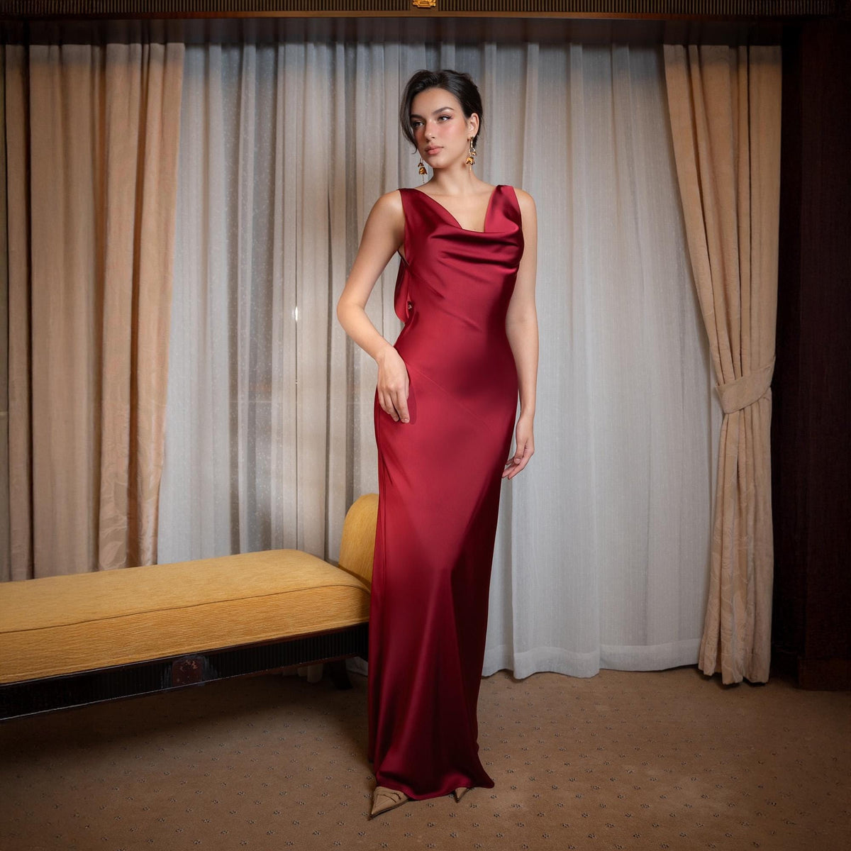 May include: A sleeveless, floor-length, burgundy-colored dress with a cowl neckline. The dress has a flowing silhouette and is made of a shiny, silky material. The model is wearing gold earrings. The background features curtains and a yellow bench.