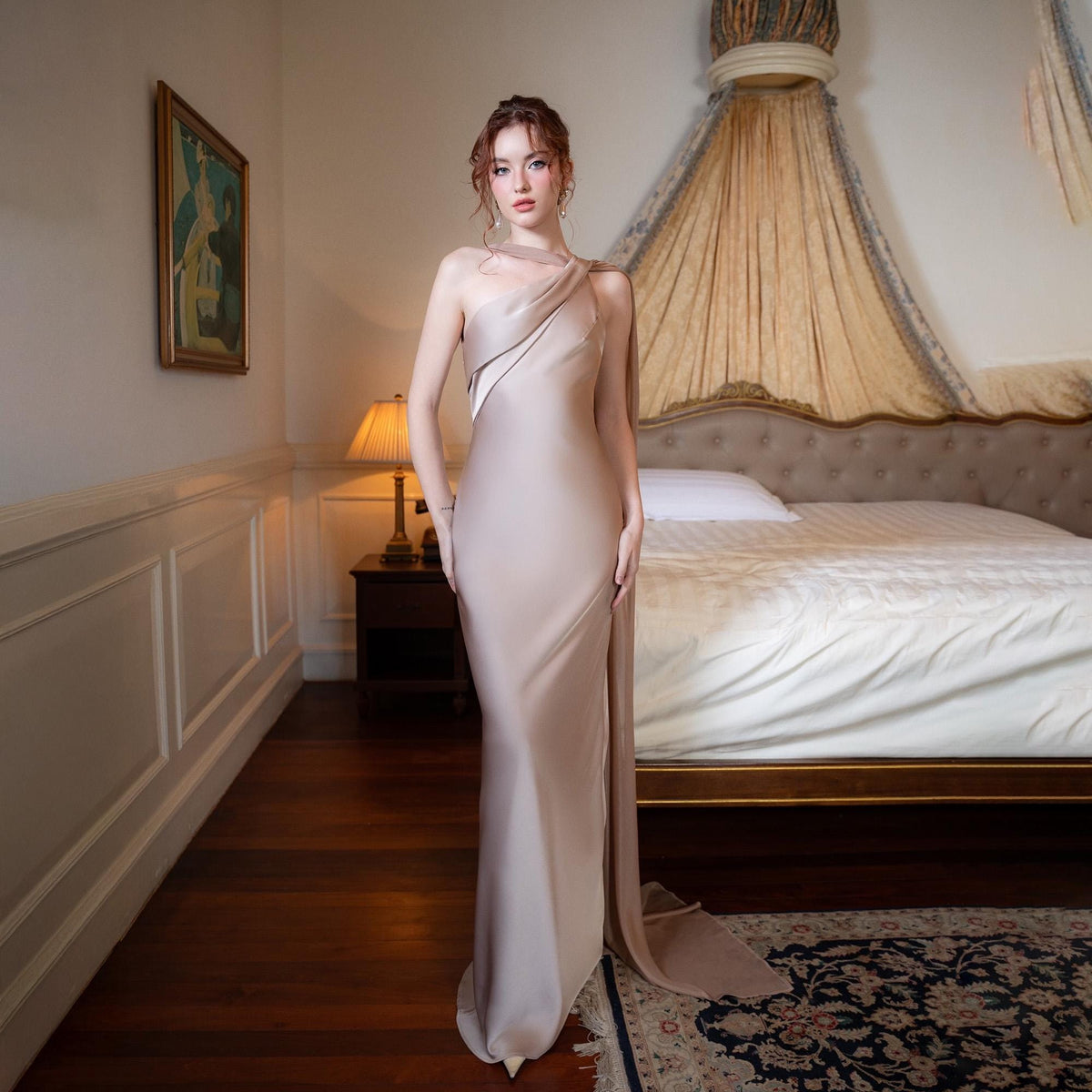 May include: A beige, floor-length gown with a draped, asymmetrical neckline and a high slit. The dress features a flowing train and is worn by a person in a bedroom setting. The room has a bed, a lamp, and a painting on the wall.
