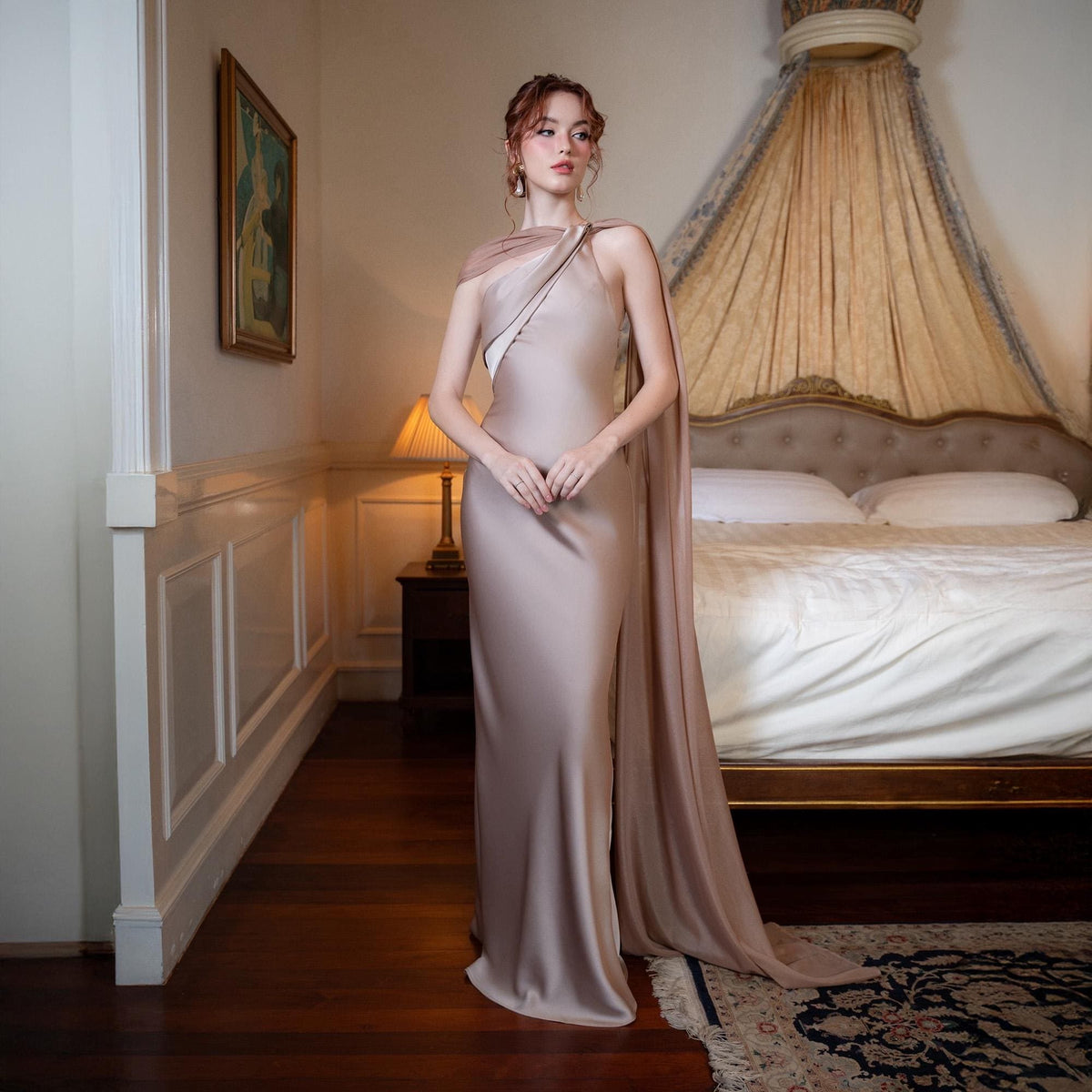 May include: A floor-length, champagne-colored gown with a draped shoulder detail and a high slit. The dress has a flowing cape-like overlay. The model is standing in a bedroom with a bed and a painting on the wall.