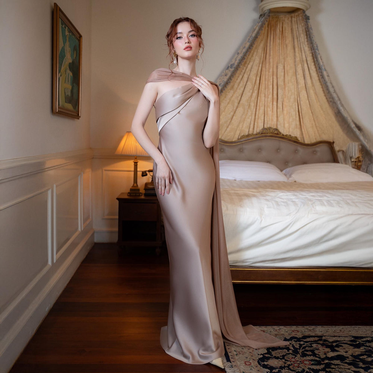 May include: A floor-length, champagne-colored gown with a draped shoulder detail and a flowing train. The dress has a fitted silhouette and a subtle sheen. The model is standing in a bedroom with a bed and a nightstand.