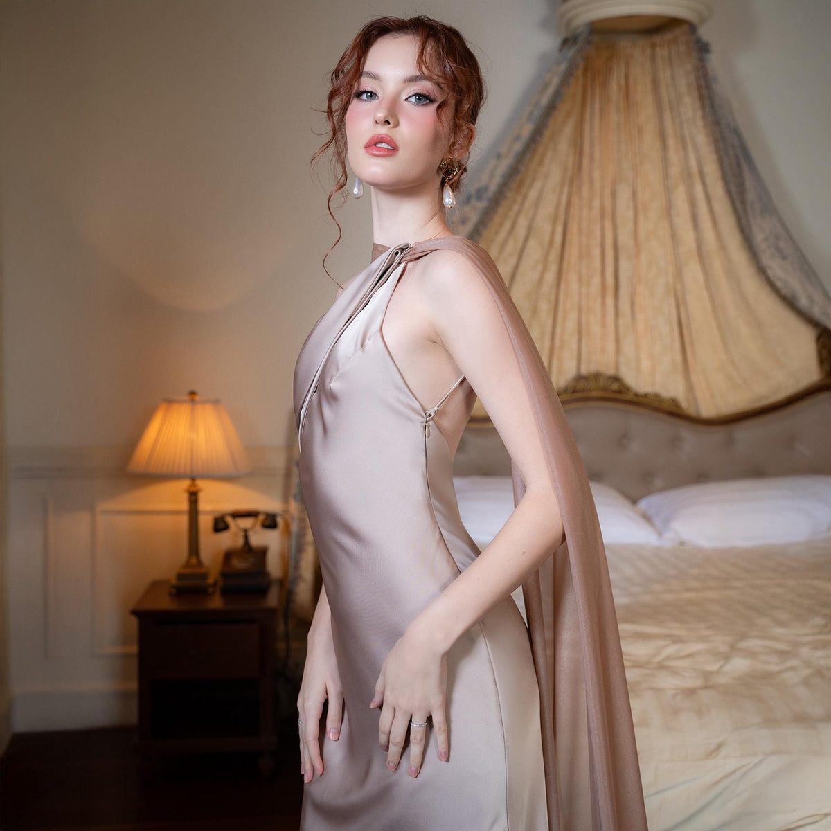 May include: A champagne-colored, floor-length gown with a draped cape detail. The dress features a halter neckline and a fitted silhouette. The model is wearing pearl earrings. The background includes a bed with a canopy and a bedside table with a lamp.