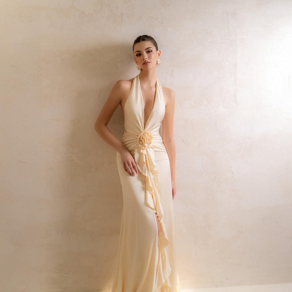May include: Ivory halter-neck gown with a plunging neckline and a flowing skirt. The dress features a floral embellishment at the waist and a ruffled detail on the side. The dress is floor-length and made of a soft, draping fabric.