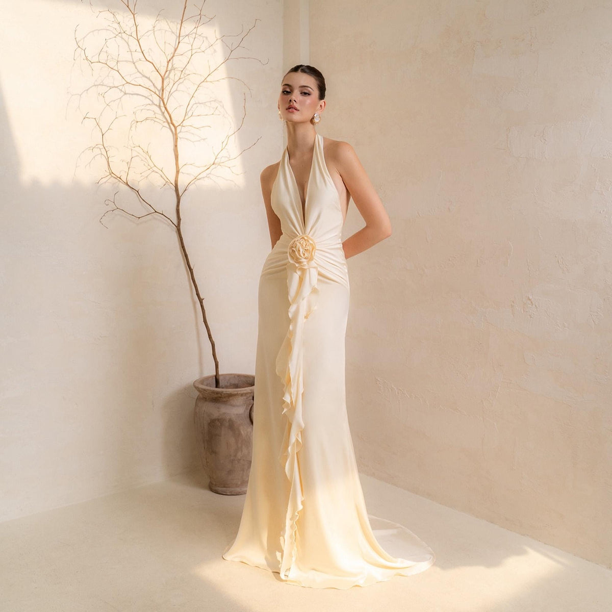 May include: Ivory halter neck gown with a plunging neckline and a floral embellishment at the waist. The dress features a flowing, floor-length skirt and ruffled detailing down the front. A classic, elegant design for special occasions.