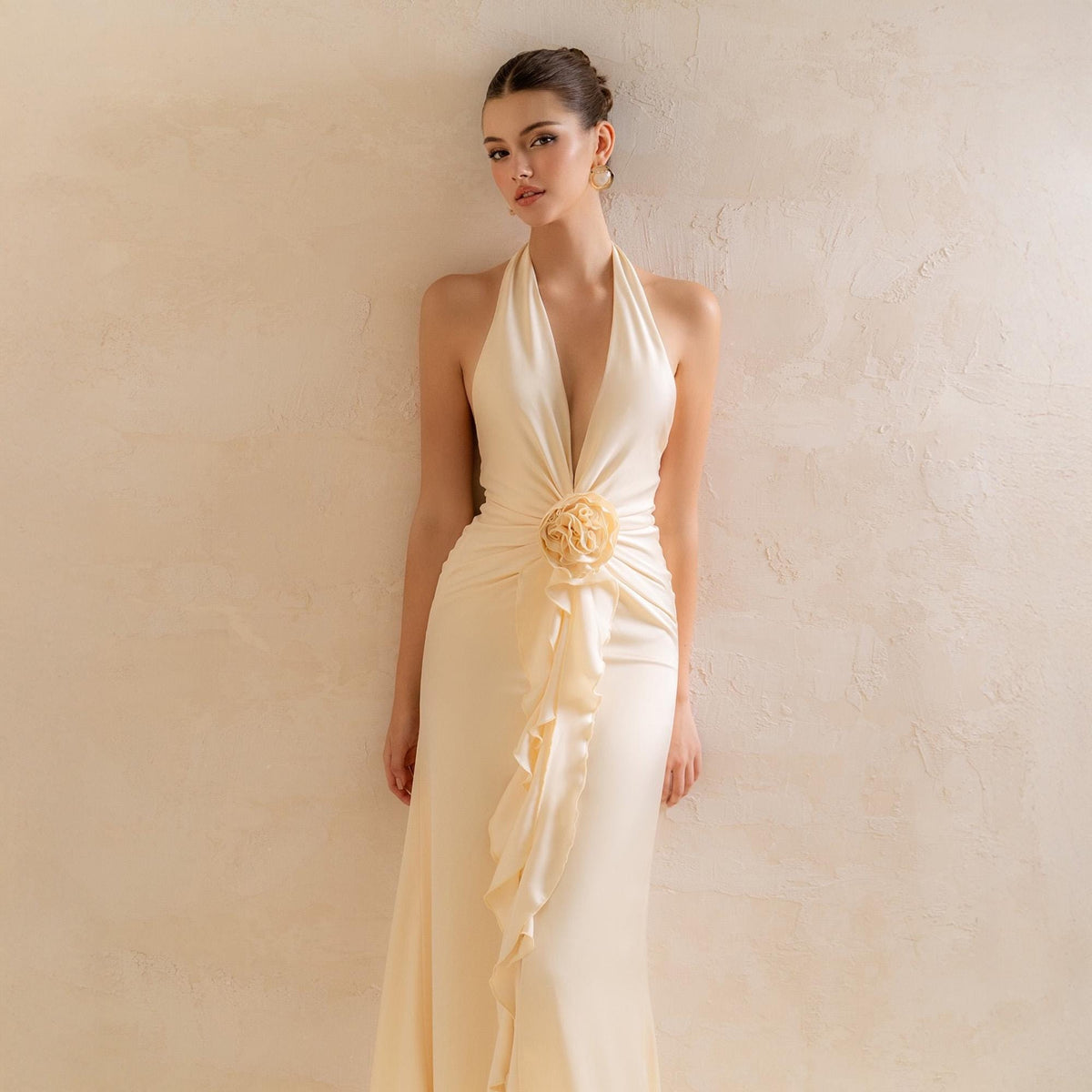 May include: Ivory halter neck gown with a plunging neckline and a fitted silhouette. The dress features a floral embellishment at the waist and a flowing ruffle detail down the front. The elegant design is suitable for formal events.